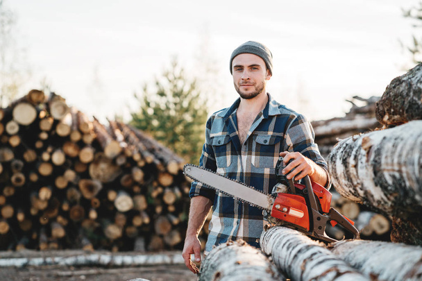 Bearded strong lumberjack wearing plaid shirt hold in hand chainsaw for work on sawmill - Фото, изображение