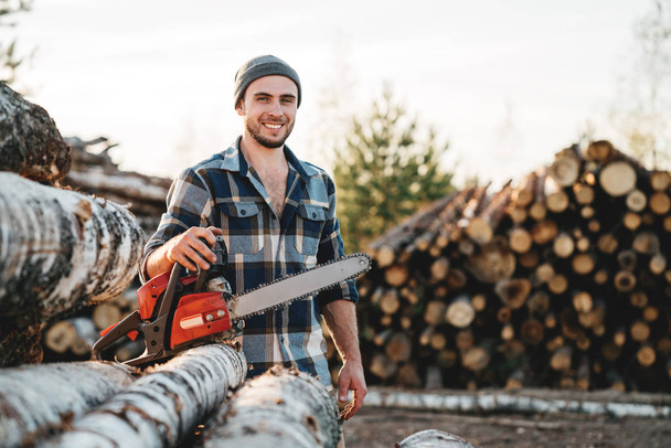 Strong bearded lumberjack wearing plaid shirt hold chainsaw in hand on background of sawmill and warehouse of trees - Foto, afbeelding