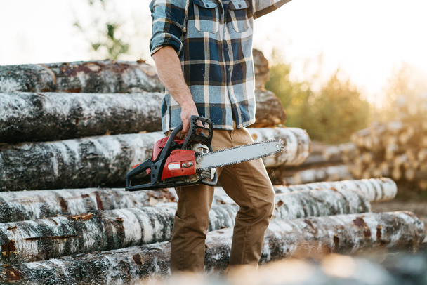 Professional bearded lumberman wearing plaid shirt hold chainsaw in hand on background of sawmill and warehouse of trees - Photo, image
