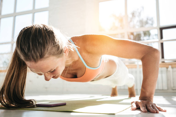 Sporty sunburn woman doing push-ups on fitness mat and using smartphone for counts results in fitness app. Female athlete exercising push up in sunny gym. Healthy lifestyle concept - Foto, Imagen