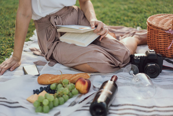 abgeschnittene horizontale Bild von Mädchen Beine beim Picknick auf einem Gras im Park mit leeren Flasche Wein und Häppchen und einer Fotokamera und ein Buch lesen. Blogger fotografiert ein Picknick - Foto, Bild