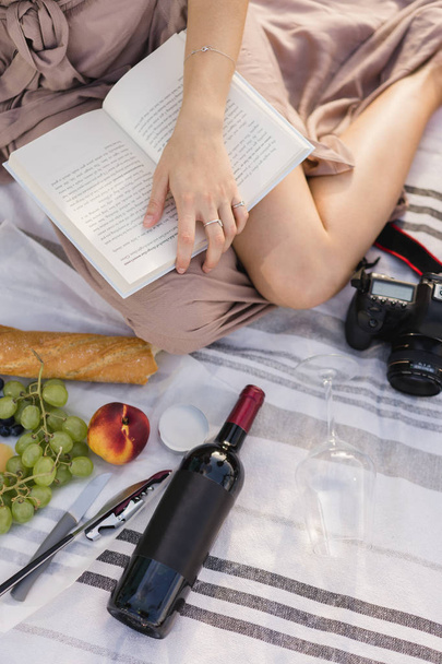 Bijgesneden afbeelding van meisjes benen lezen van een boek terwijl het hebben van een romantische picknick zitten in een stad Parc en drinken van wijn met fruit. Mockup voor een fles rode wijn met een ruimte voor uw logo of ontwerp - Foto, afbeelding