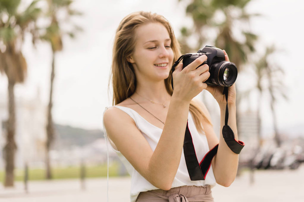 Pretty young girl laughing while taking pictures on professional dslr camera walking outdoors in the park on the summer sunny day, traveling, making photos, photography as a hobby, looking into camera - Foto, afbeelding