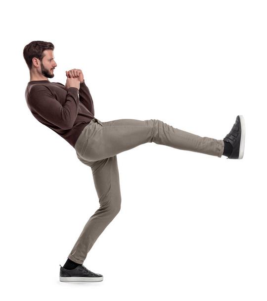 An isolated bearded man in casual clothes stands on white background in the middle of leg kick. - Foto, afbeelding