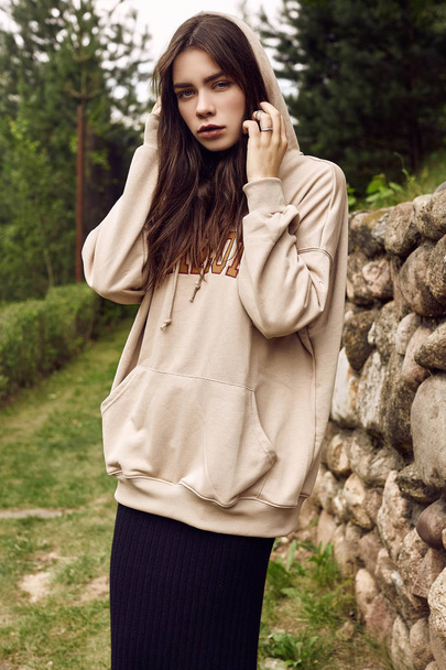 Portrait of gorgeous brunette woman in fashion hoodie posing in the garden alley - Photo, Image