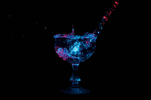 Water wordt gegoten in een glas goblet en spetterend onder gekleurde lampjes op een zwarte achtergrond - Foto, afbeelding