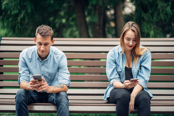Phone addicted couple on the bench in park. Man and woman using their smartphones, addiction - 写真・画像