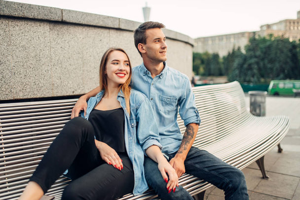 Young love couple sitting on the bench in summer city park. Smiling teenagers poses together, cityscape on background - Photo, Image