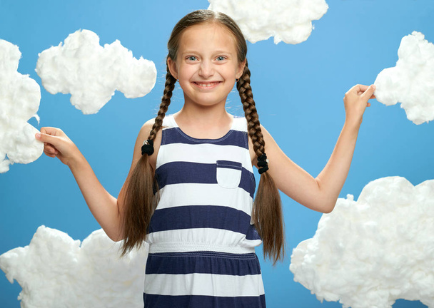 girl dressed in striped dress posing on a blue background with cotton clouds, the concept of summer, holiday and happiness - Photo, Image