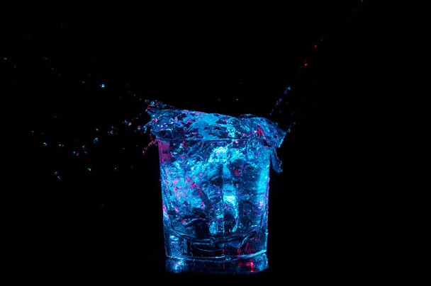 Water splashing out from a full rocks tumbler in bright blue light with red accents isolated on a black background - Photo, Image