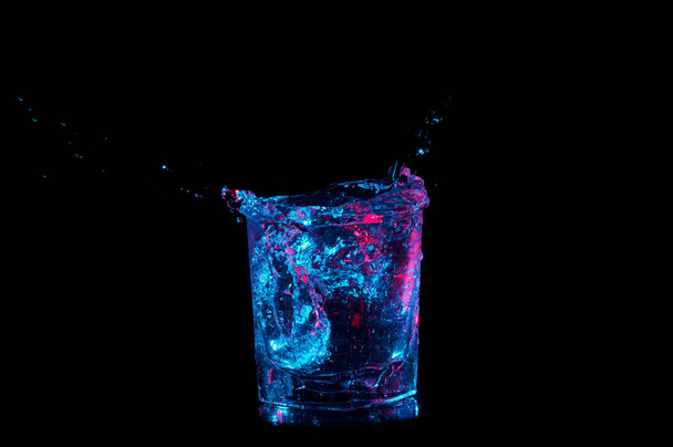 Flüssigkeit spritzt sanft aus einem Steinglas in blauem Licht auf schwarzem Hintergrund - Foto, Bild