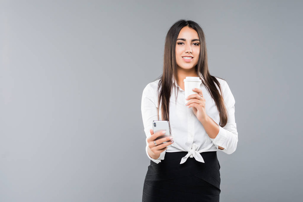 Portrait of a satisfied young business woman using mobile phone while holding cup of coffee to go isolated over white background - Foto, afbeelding