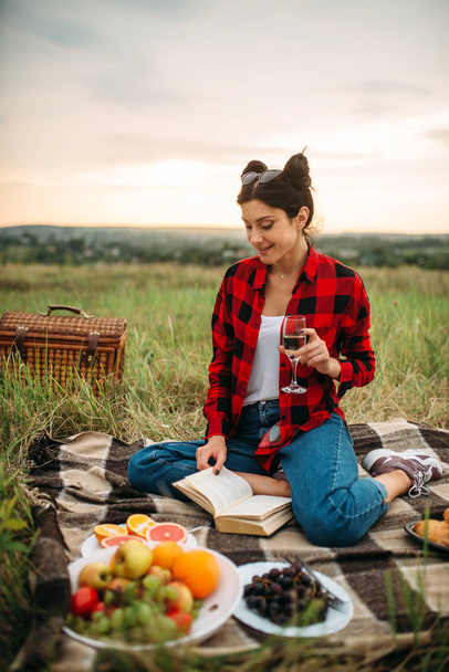 Cute woman with glass of wine reads book, picnic on meadow. Romantic junket, happy holiday - Fotoğraf, Görsel