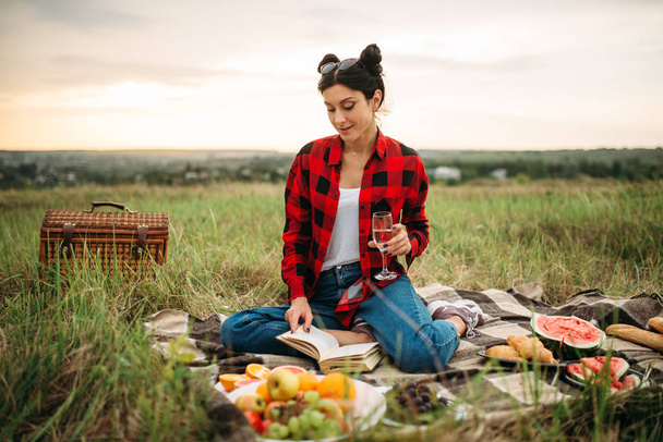 Cute woman with glass of wine reads book, picnic on meadow. Romantic junket, happy holiday - Photo, image