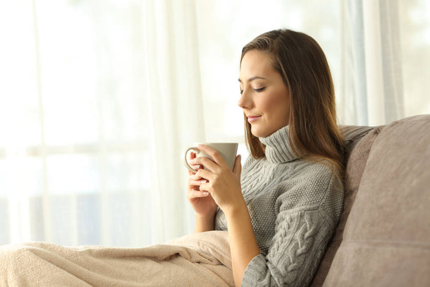 Pensive lady holding a cup of coffee in winter sitting on a couch in the living room at home - Foto, immagini