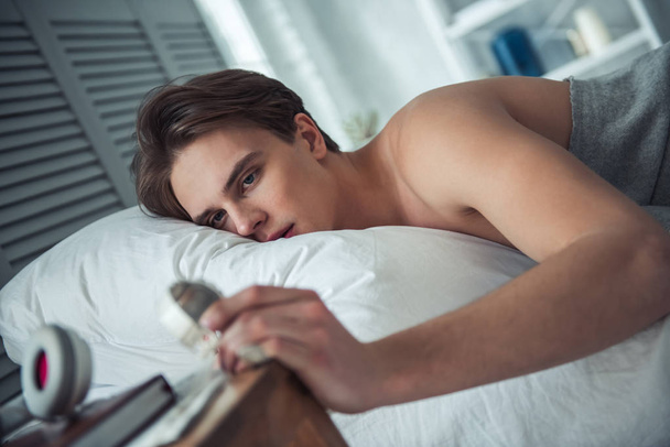 Komea nuori mies katsoo kelloaan makatessaan sängyssään kotona aamulla.
 - Valokuva, kuva