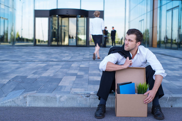 Fired business man sitting frustrated and upset on the street near office building with box of his belongings. He lost work - 写真・画像