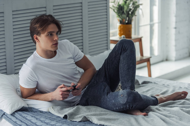Beau jeune homme en vêtements décontractés joue console de jeu vidéo tout en étant assis sur son lit à la maison
 - Photo, image