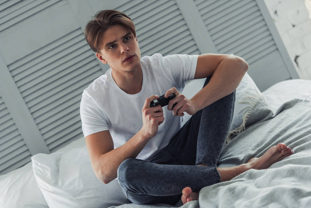 Handsome young man in casual clothes is playing video game console while sitting on his bed at home - Фото, изображение