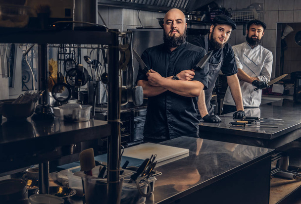 Team of professional bearded cooks dressed in uniforms posing with knives in kitchen. - Fotó, kép