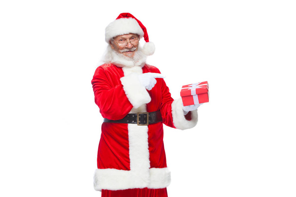 Christmas. Smiling Santa Claus in white gloves is holding a gift red box with a bow. Pointing at the gift. Isolated on white background. - Фото, изображение
