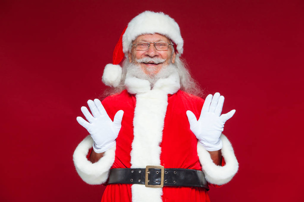 Christmas. Good Santa Claus in white gloves shows faces, grimaces, shows his tongue. Not standard behavior. Isolated on red background. - Foto, Bild