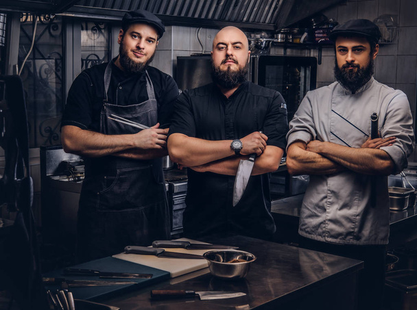 Team of professional bearded cooks dressed in uniforms posing with knives in kitchen. - Photo, Image