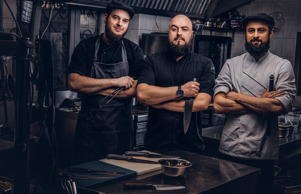 Team of professional bearded cooks dressed in uniforms posing with knives in kitchen. - Foto, afbeelding
