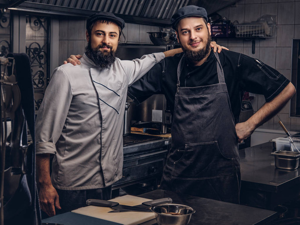 Two bearded cooks friend dressed in uniforms and hats embrace while posing in kitchen. - Photo, Image