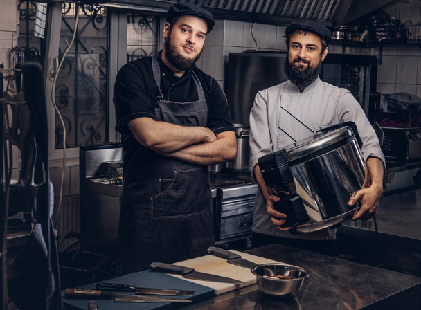 Дві жорстокі кухарі, одягнені в уніформу і капелюхи, стоять з великим мультиваркою на кухні
. - Фото, зображення