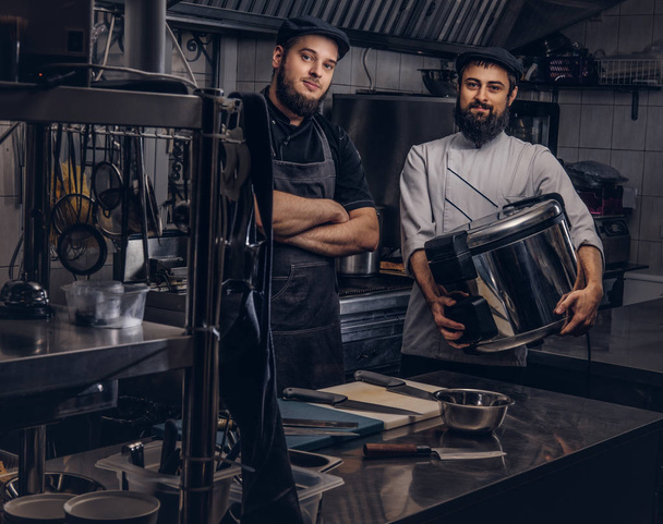 Дві жорстокі кухарі, одягнені в уніформу і капелюхи, стоять з великим мультиваркою на кухні
. - Фото, зображення