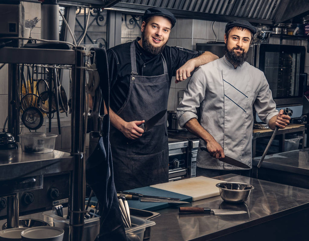 Δύο μάγειρες γενειοφόρος ντυμένοι με στολές και καπέλα ποζάρει στην κουζίνα, βλέπουν μια φωτογραφική μηχανή. - Φωτογραφία, εικόνα