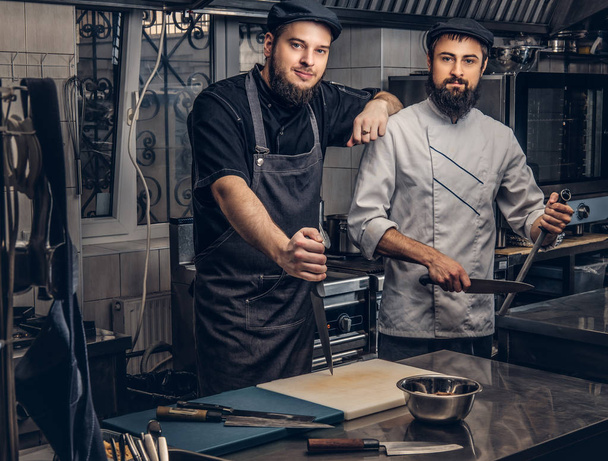Deux cuisiniers barbus vêtus d'uniformes et chapeaux posant dans la cuisine, regardant une caméra
. - Photo, image