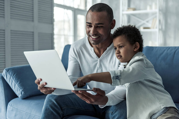 Счастливые афро-американский отец и сын в повседневной одежде используют ноутбук, сидя на диване дома, папа улыбается
 - Фото, изображение