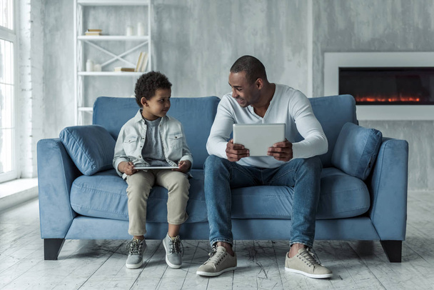 Boldog Afro-amerikai apa és fia, alkalmi ruhák használ digitális tabletta, és beszél, miközben otthon a fotelben ülve, apa mosolyogva - Fotó, kép