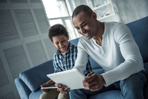 Feliz pai e filho afro-americano em roupas casuais estão usando tablets digitais e sorrindo enquanto estão sentados no sofá em casa
 - Foto, Imagem