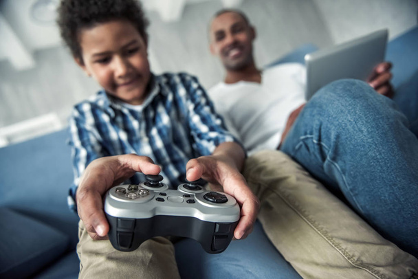 Afro American father and son in casual clothes using gadgets, sitting on couch at home, boy is playing video game console in focus - Photo, Image