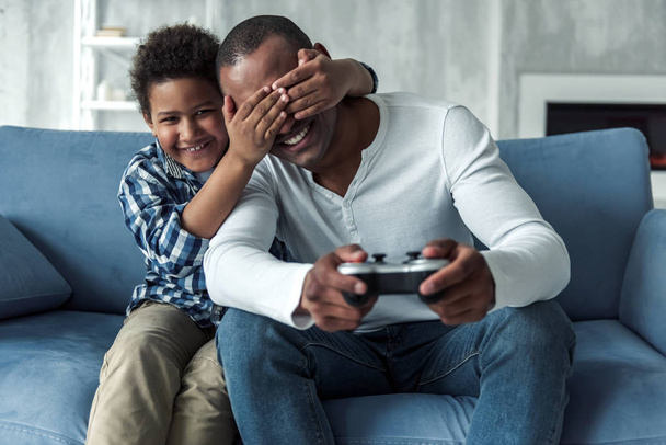 Happy Afro American father and son on couch at home, boy is covering his dad's eyes and smiling while dad is playing video game console - Photo, Image