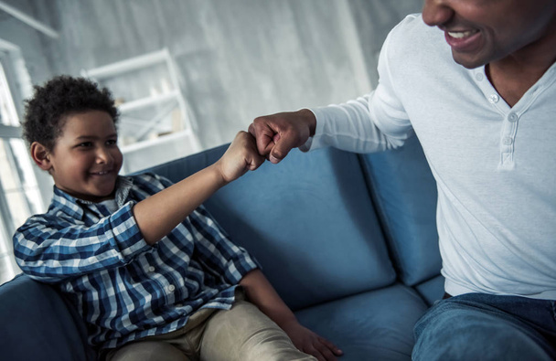 Happy Afro American father and son in casual clothes are doing a fist bump and smiling while sitting on couch at home - Photo, Image