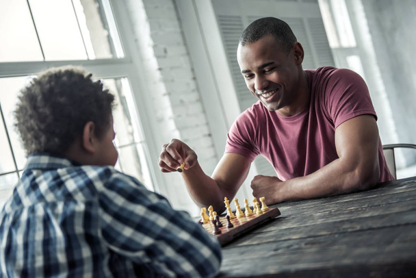 Afro American father and son in casual clothes are playing chess while spending time together at home, man is smiling - Foto, Bild