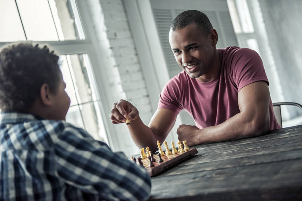 Afro-americano pai e filho em roupas casuais estão jogando xadrez enquanto passam o tempo juntos em casa, o homem está sorrindo
 - Foto, Imagem