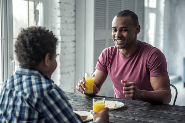 アフロ ・ アメリカ人の父親とカジュアルな服装の息子は、木製のテーブルで一緒に家で食べる、男は笑みを浮かべて - 写真・画像