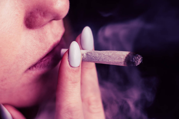 cannabis weed, a joint in his hands A woman smokes . Smoke on a black background. Concepts of medical marijuana use and legalization of the cannabis. On a black background Pink tinting - 写真・画像