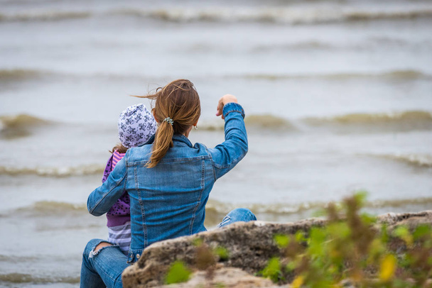 Widok z tyłu nowoczesnej matki i dziecka w kolorowe ubrania na plaży rzucanie kamieniami w seashore. - Zdjęcie, obraz