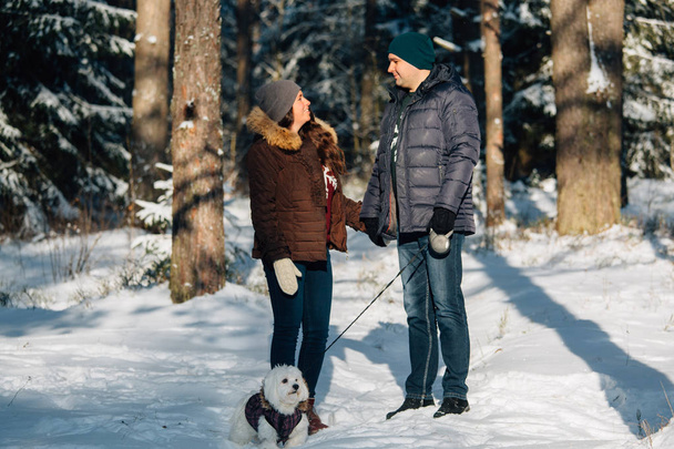 Cute young couple having fun in winter forest with their pretty little white dog. Man and woman in sweater with a snowflake - Fotoğraf, Görsel