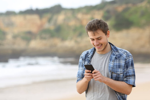 Happy teen guy using a smart phone walking on the beach - 写真・画像