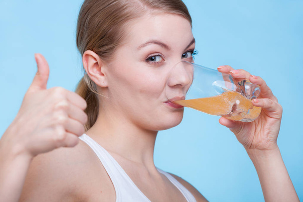 Food, health concept. Woman holding glass of orange flavored drink and drinking from it showing thumb up gesture. Studio shot on blue background - Zdjęcie, obraz