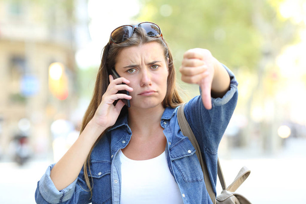 Front view portrait of an angry girl talking on phone with thumbs down in the street - Фото, зображення