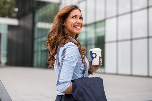 Hermosa joven empresaria sosteniendo un café para ir frente a un edificio de oficinas durante el día
 - Foto, imagen