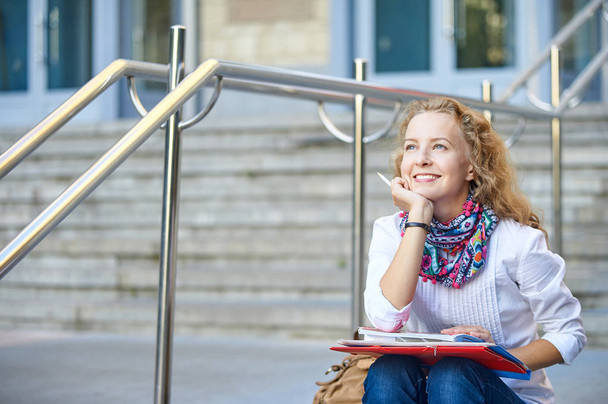 Young smiling student woman writing and reading book on stairs of university or college, preparing for exams. Education concept, copy space - Foto, imagen